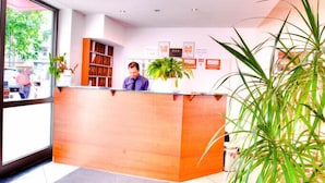 A reception area with a wooden desk, a receptionist, a large potted plant, and a tiled floor. - Hotel Himalaya Frankfurt City Messe (Frankfurt)