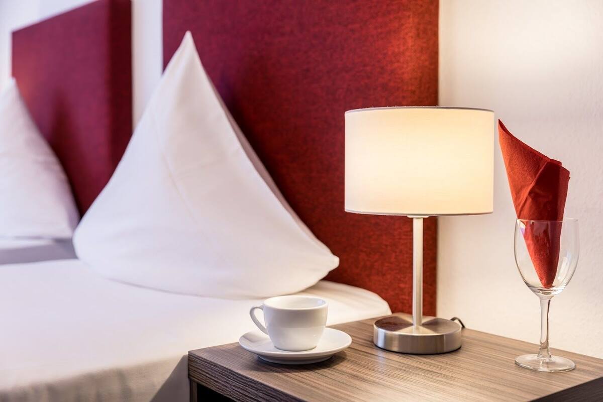 A neatly made hotel room with a bed, a bedside table with a lamp, a cup, and a glass.