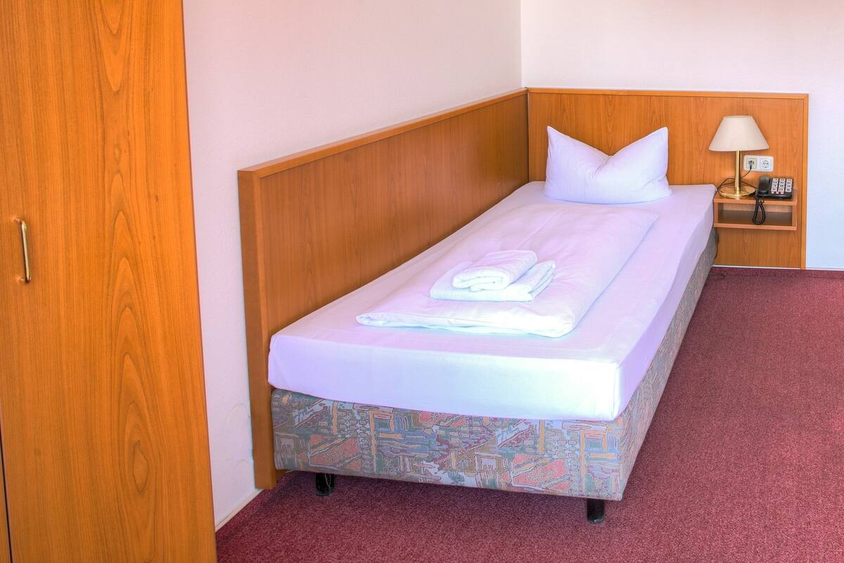 A single bed with a patterned mattress, a wooden headboard, a bedside table with a lamp, and a red carpeted floor.