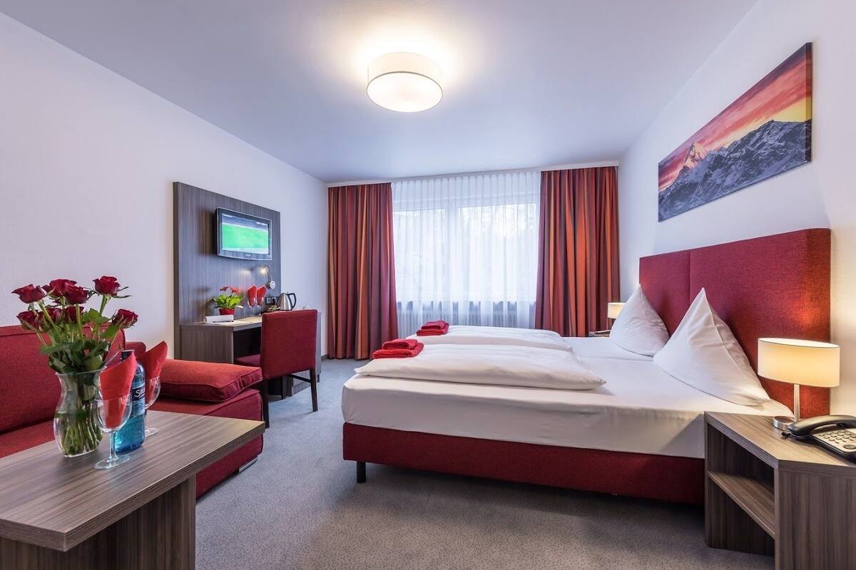 A hotel room with a bed, a desk, a TV, a red sofa, and a painting on the wall.