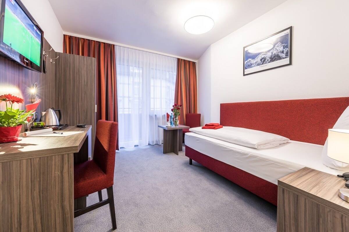 A hotel room with a bed, a desk, a TV, a chair, a small table, and a window with curtains.