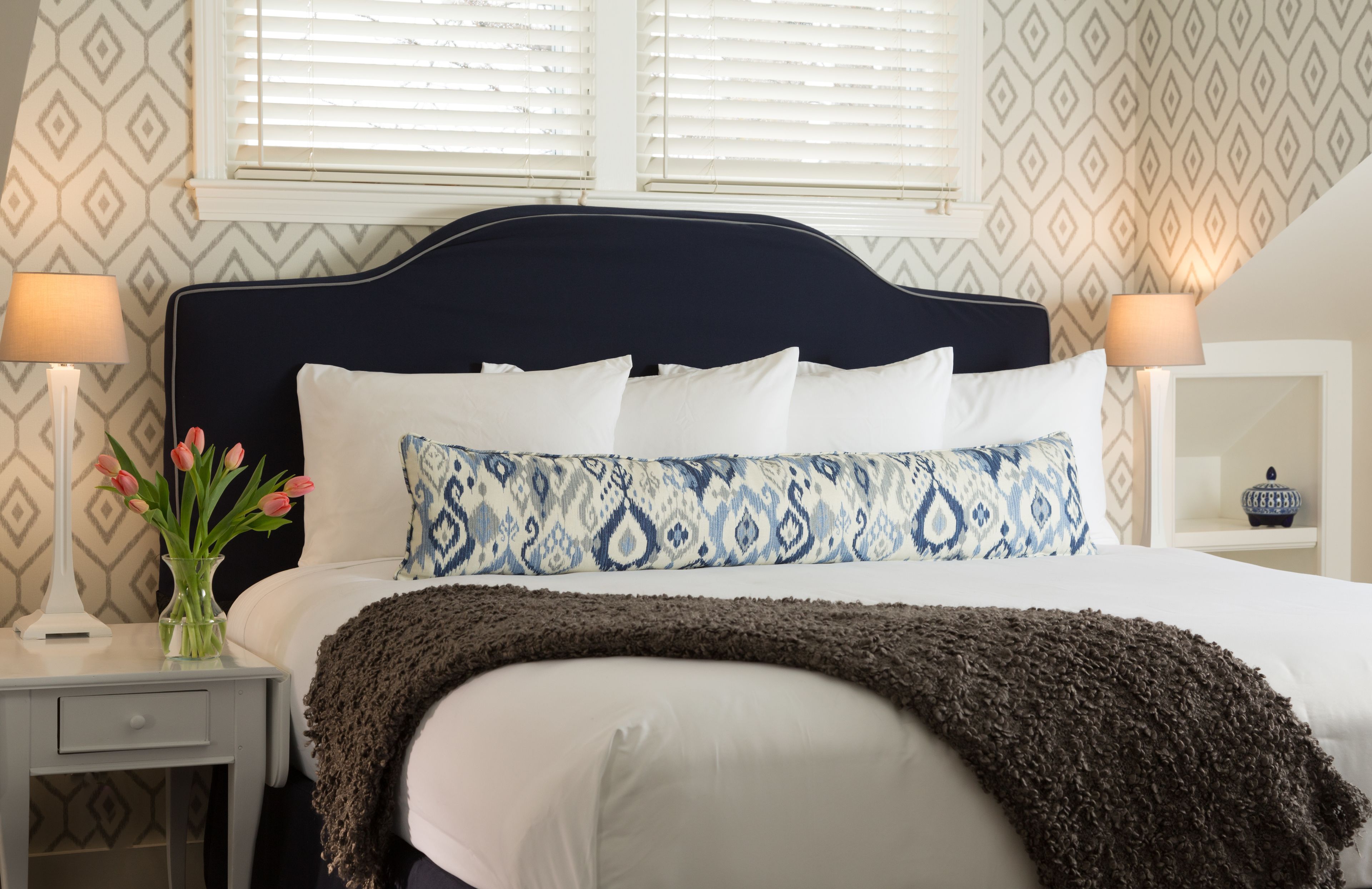 A neatly made bed with a dark headboard, white pillows, and a patterned throw blanket. There are two bedside lamps and a vase with flowers on a nightstand.