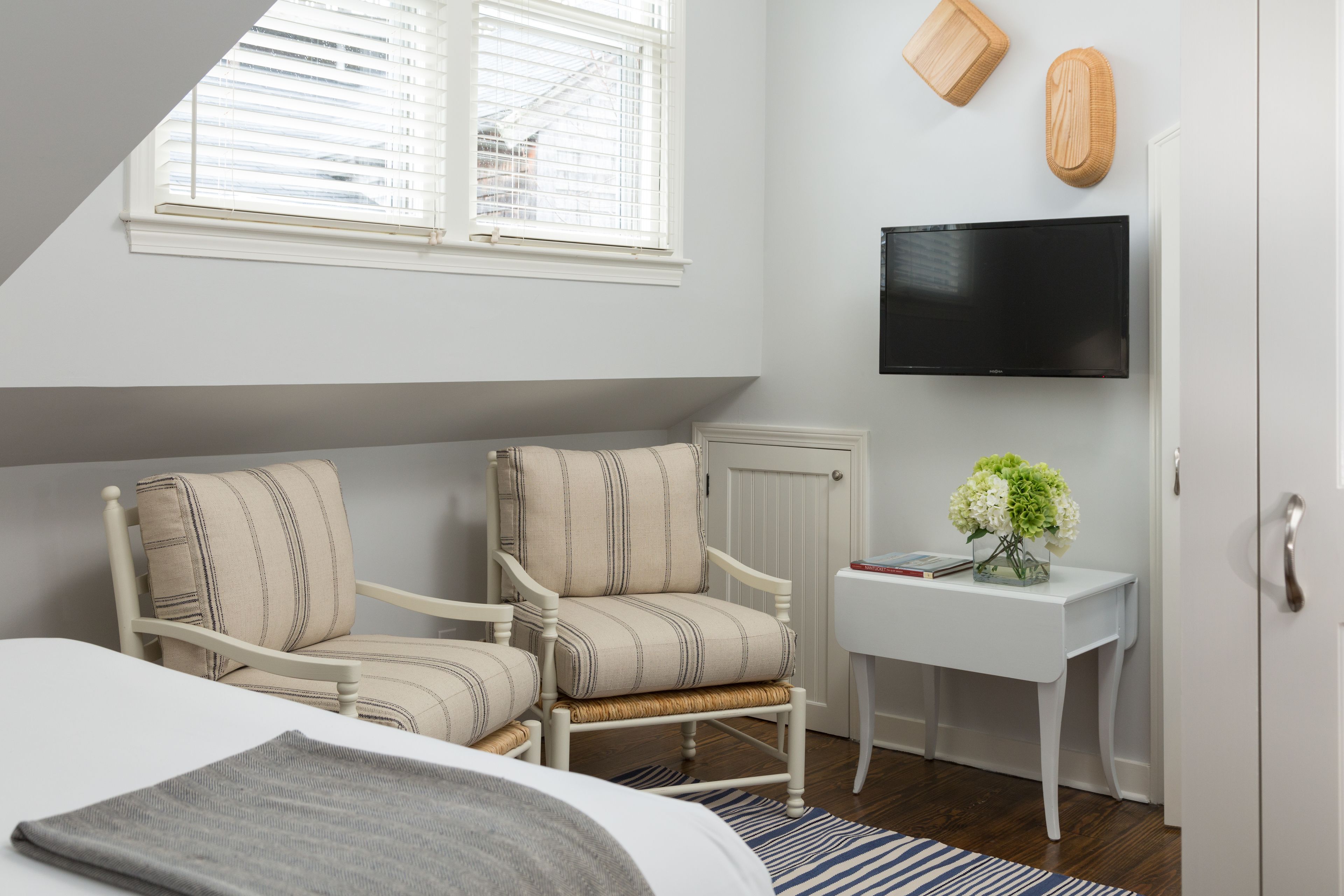 A bedroom with a bed, two upholstered chairs, a small white table with a vase of flowers, a mounted TV, and a window with blinds.