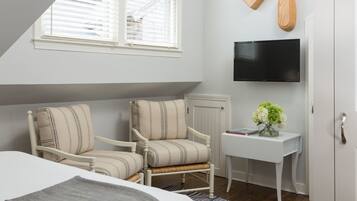 A bedroom with a bed, two upholstered chairs, a small white table with a vase of flowers, a mounted TV, and a window with blinds.