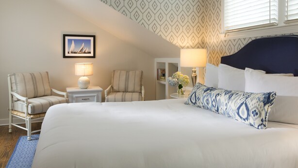 A hotel room with a large bed, two armchairs, a nightstand with a lamp, a wall-mounted picture, and a patterned wallpaper. - Brass Lantern Inn (Nantucket)