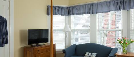 Un dormitorio con cama, televisor sobre un aparador de madera, sofá azul, ventana con persianas y una lámpara.