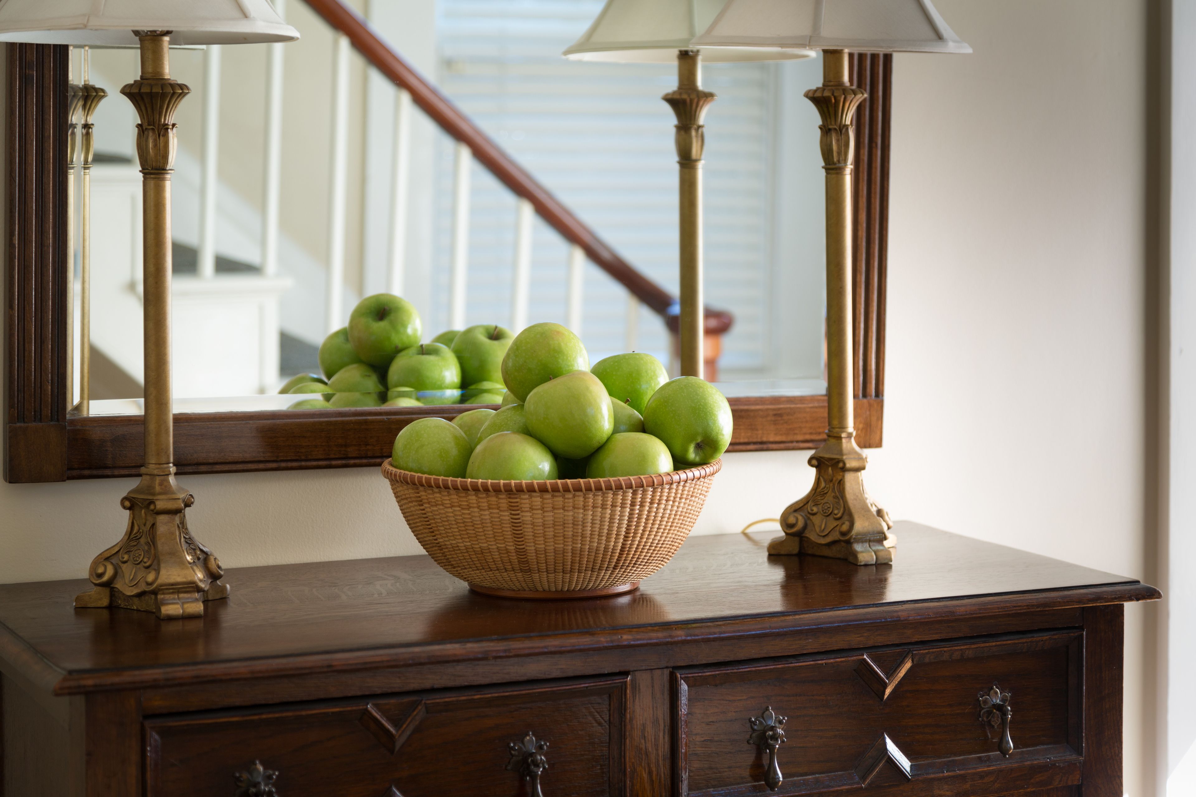Een houten dressoir met een rieten schaal groene appels, twee messing lampen en een spiegel die een trap weerspiegelt.