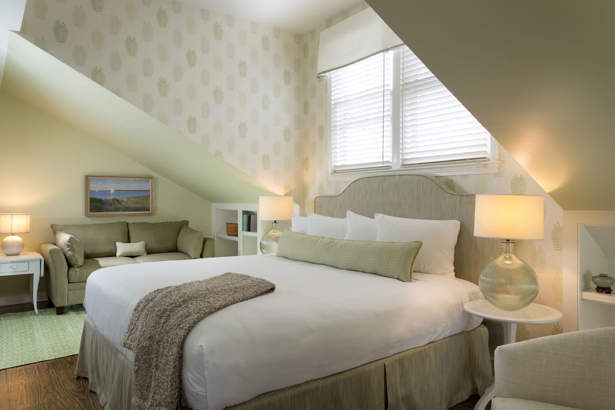 A bedroom with a large bed, a nightstand, a lamp, a chair, and a window with blinds.