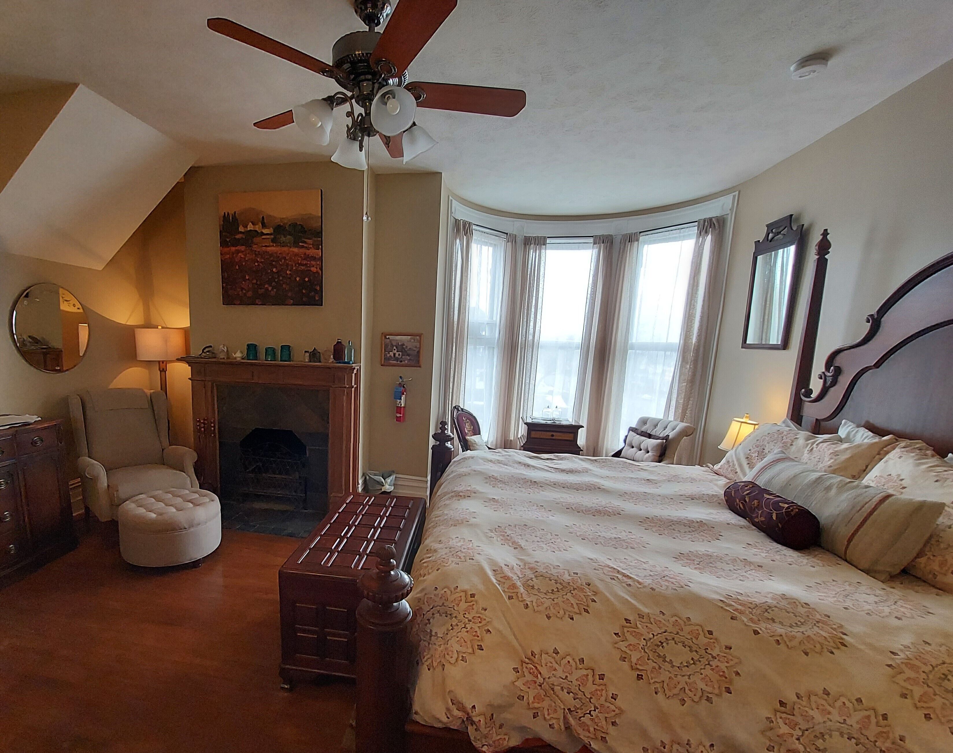 Un dormitorio con cama, una silla, chimenea, ventilador de techo y una ventana con cortinas.