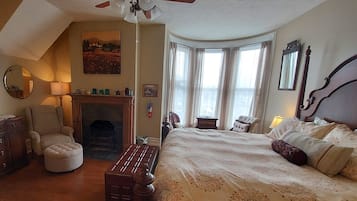 A bedroom with a bed, a chair, a fireplace, a ceiling fan, and a window with curtains.