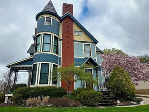 Une grande maison à deux étages, dotée d’une tourelle remarquable et d’une cheminée, entourée d’un jardin bien entretenu et d’arbres en fleurs.