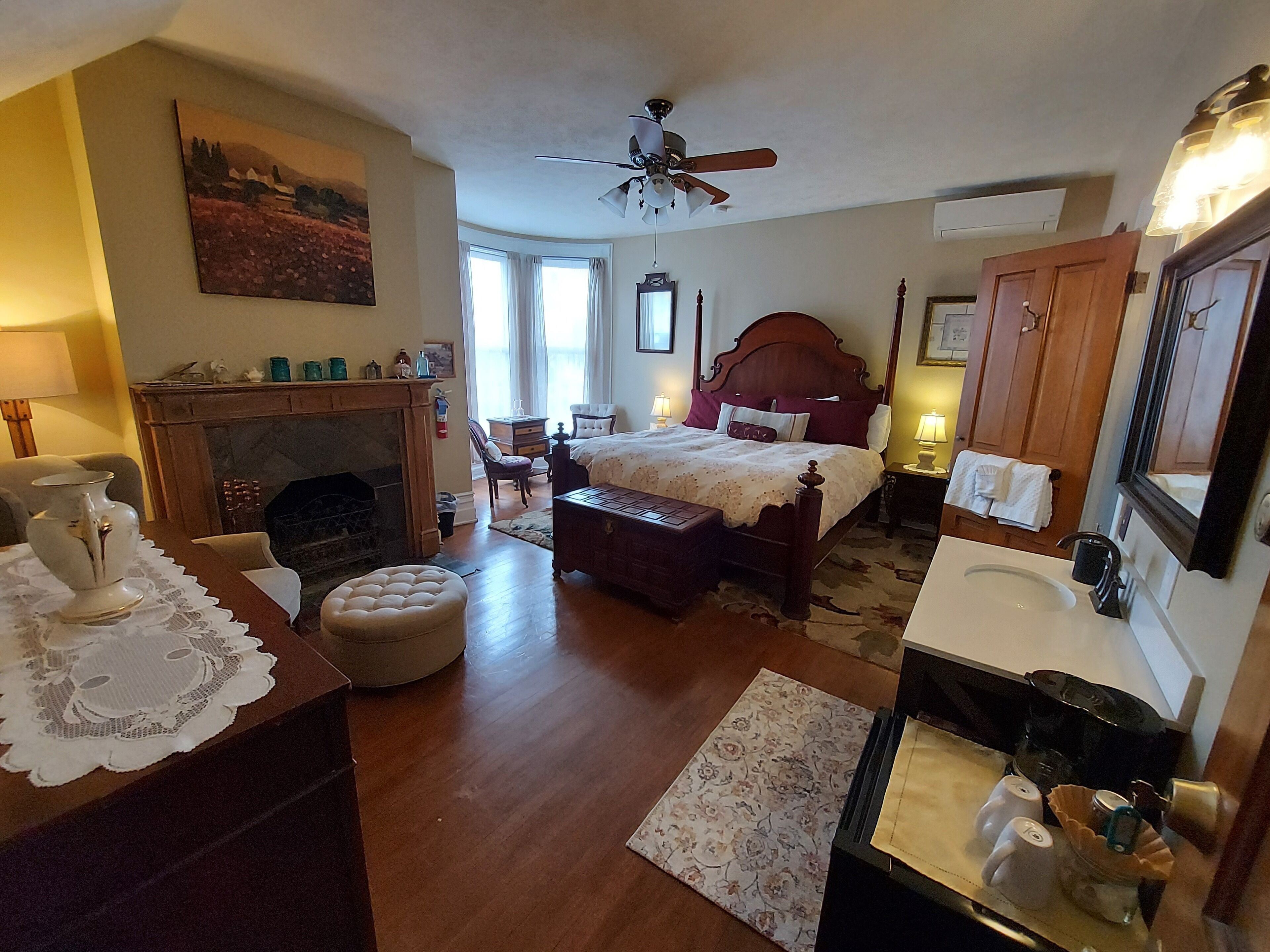 A bedroom with a wooden bed, a fireplace, a desk with a vase, and a bathroom with a sink and mirror.