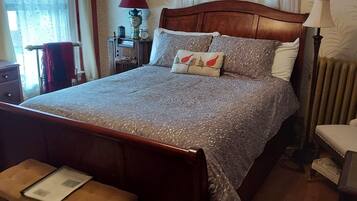 A neatly made bed with a patterned bedspread and pillows, a wooden headboard, a bedside table, and a chair with a cushion.