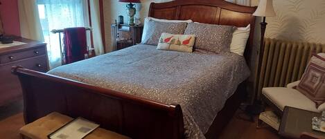 A neatly made bed with a patterned bedspread and pillows, a wooden headboard, a bedside table, and a chair with a cushion.