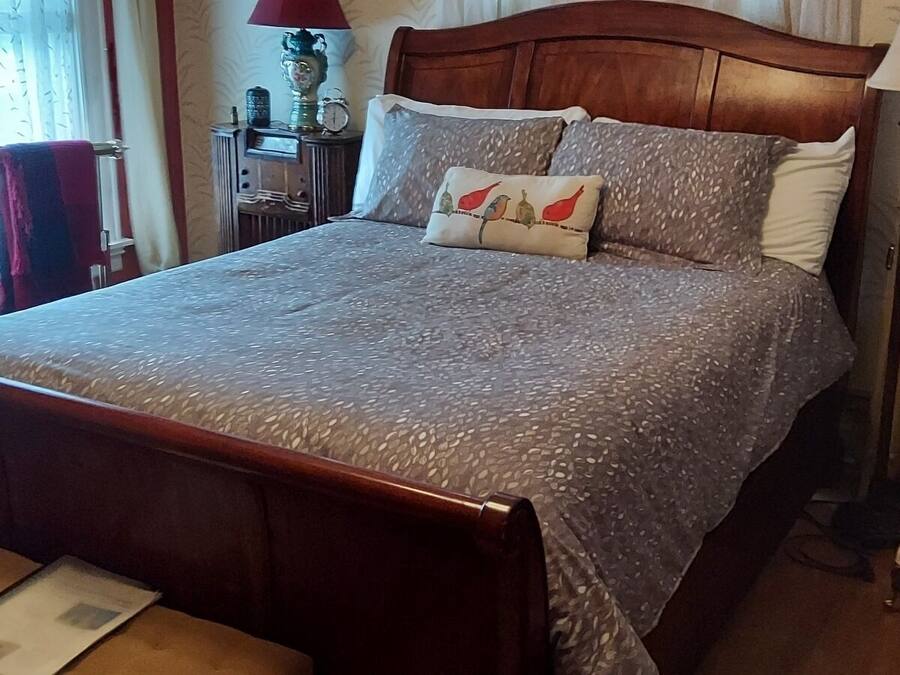 A neatly made bed with a patterned bedspread and pillows, a wooden headboard, a bedside table, and a chair with a cushion.