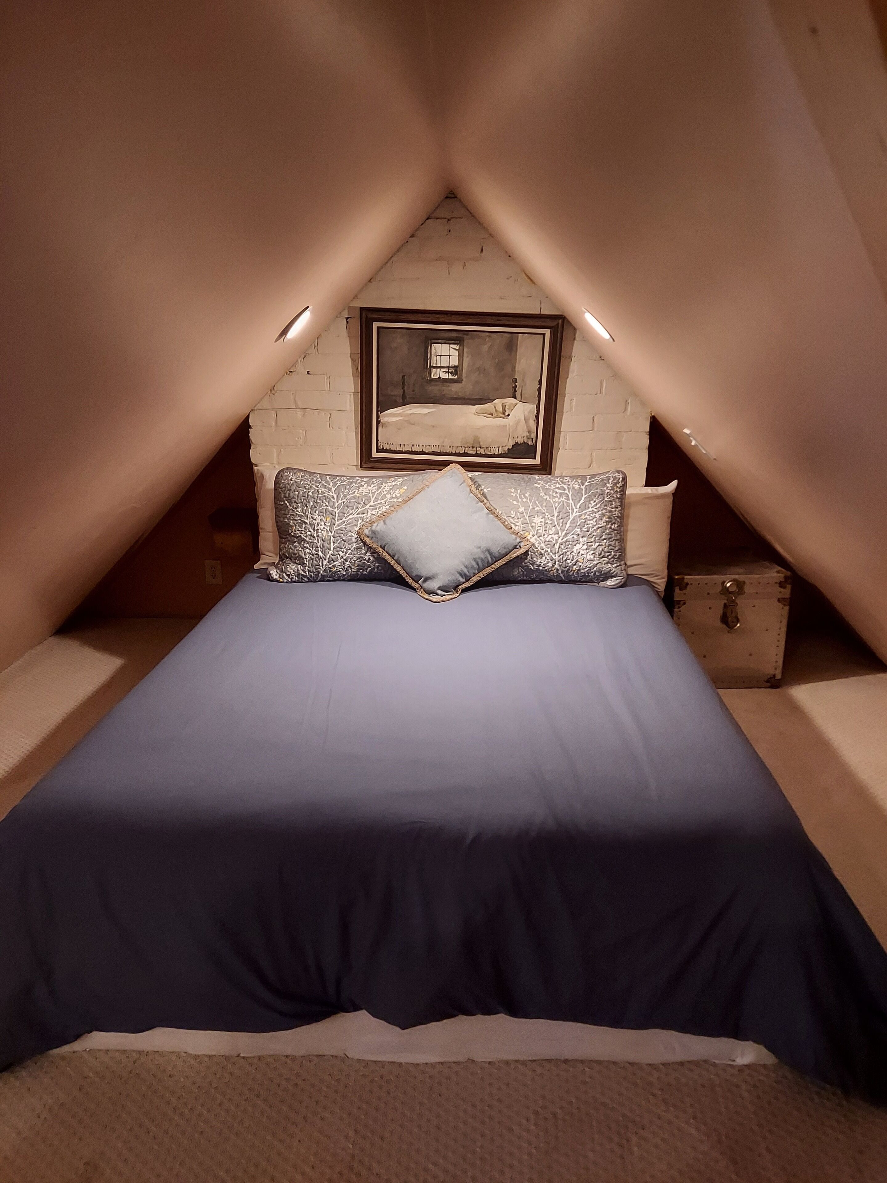 A bedroom with a sloped ceiling, a bed with a blue bedspread, two pillows, a nightstand, and a framed picture on the wall.