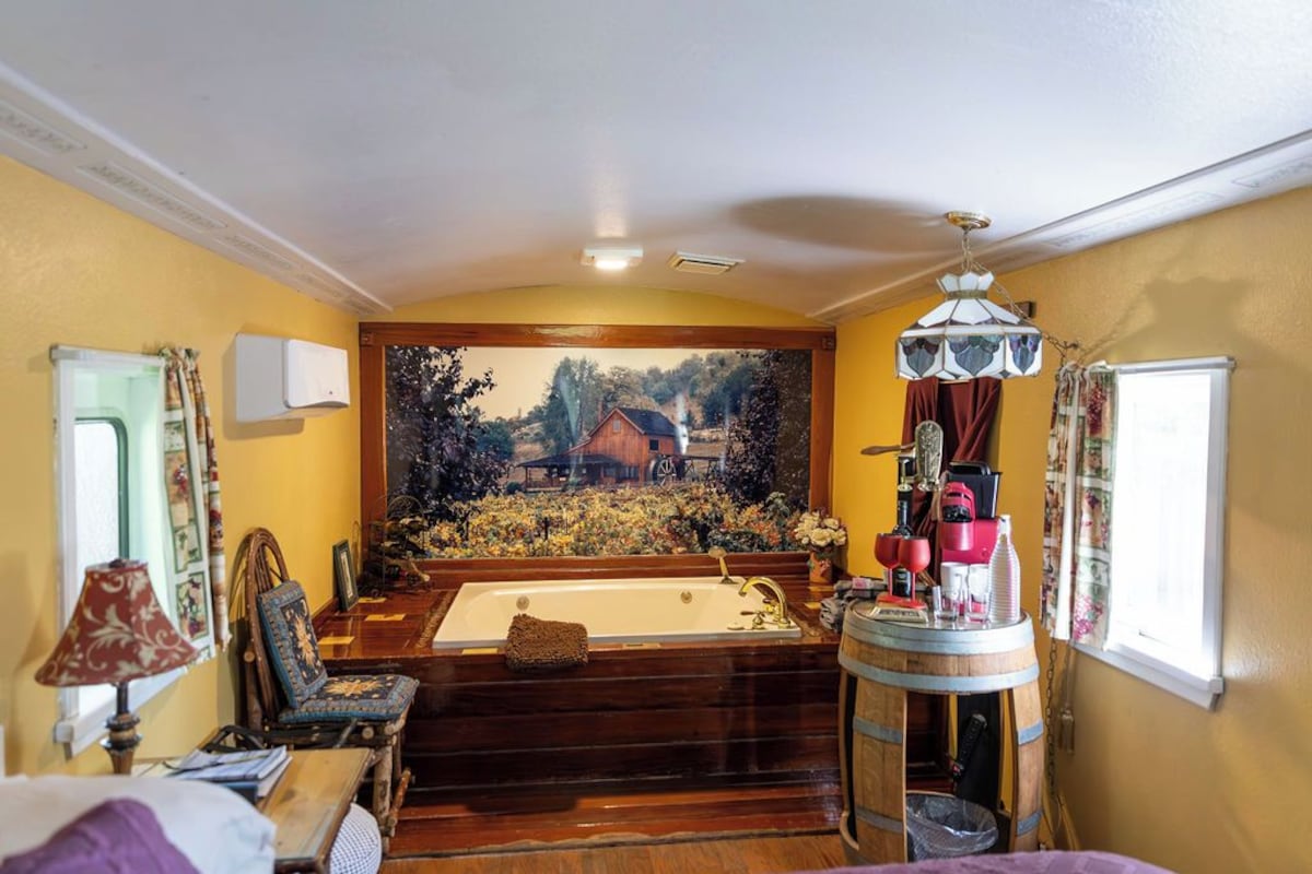 A room with a large window, a wooden bathtub, a chair, a table, and a lamp.