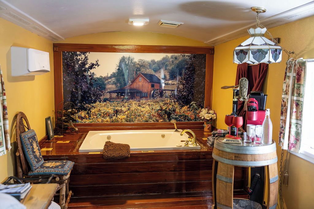 A room with a large painting of a rural scene, a bathtub, a wooden chair, and a round table with a coffee machine.