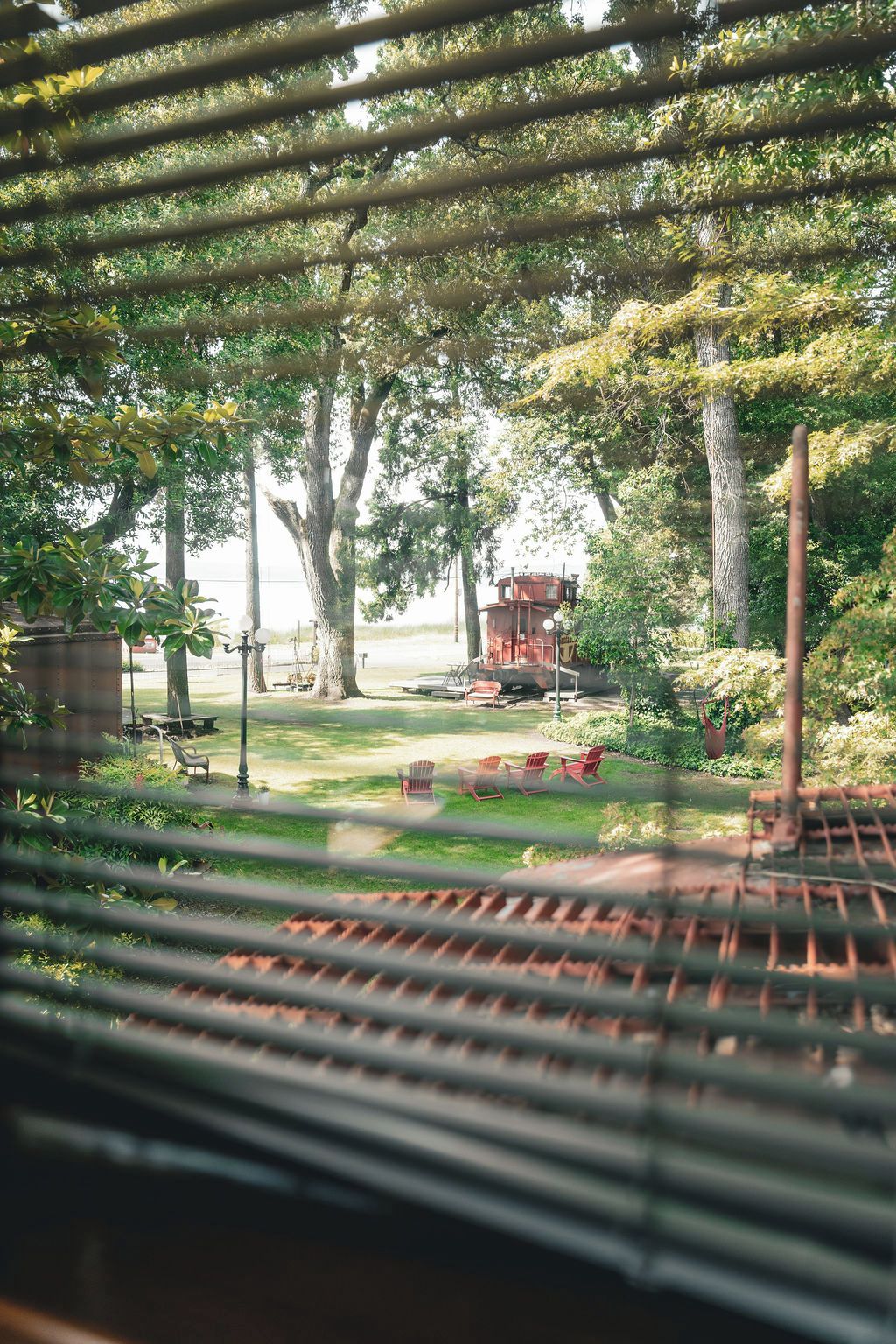 Uitzicht door een raam met jaloezieën, met een tuin, een rode constructie, mogelijk een prieel, en een picknicktafel.
