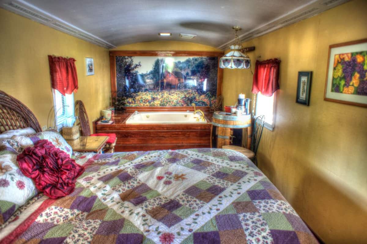 Une chambre à coucher avec un grand lit, une tête de lit en bois, une fenêtre avec des rideaux rouges, un tableau au mur et une petite table avec une lampe.