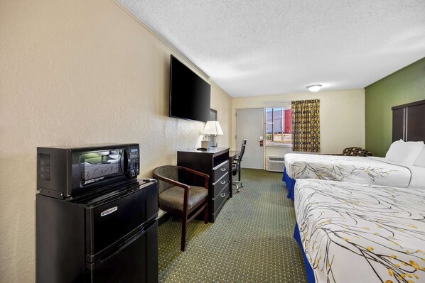 A hotel room with a flat-screen TV, a desk with a lamp, a chair, a bed with a floral bedspread, and a window with curtains. - Americas Best Value Inn Ft. Jackson (Columbia)