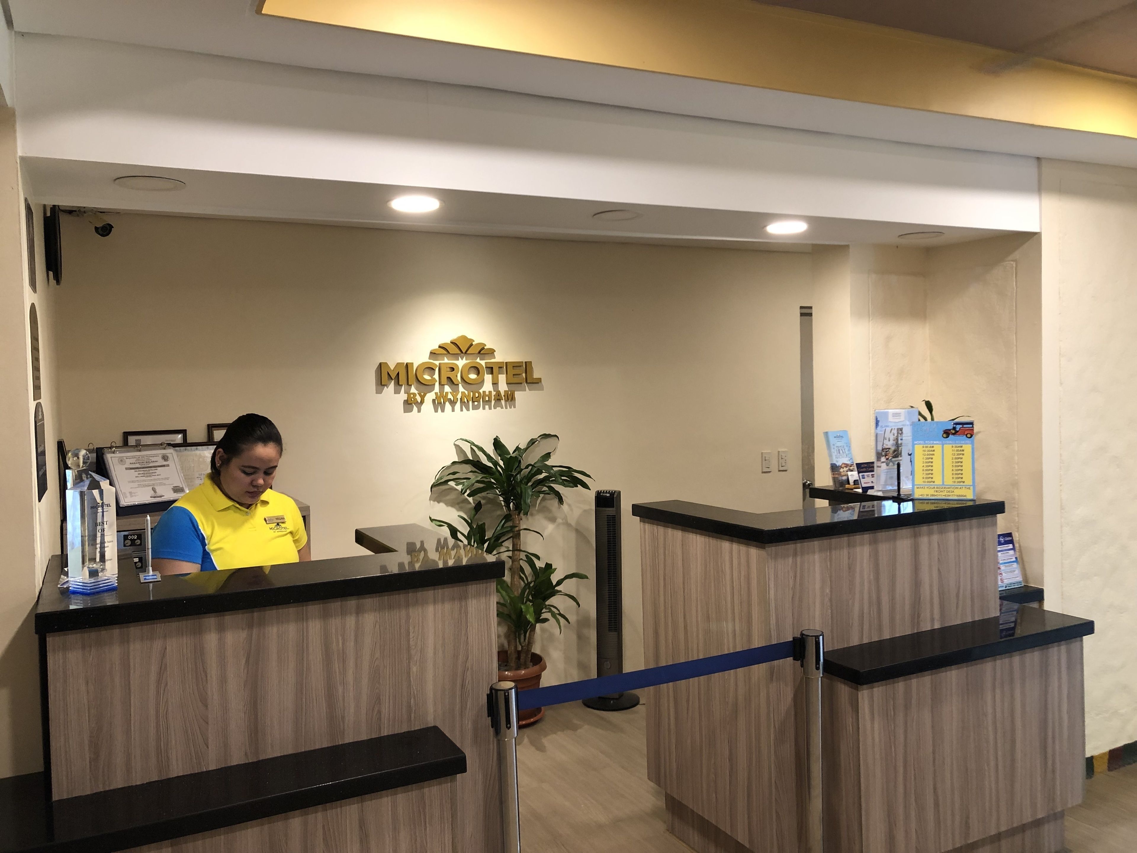 Un mostrador de recepción con una recepcionista, un cartel que dice "MICROTEL BY WYNDHAM" y una planta en maceta.