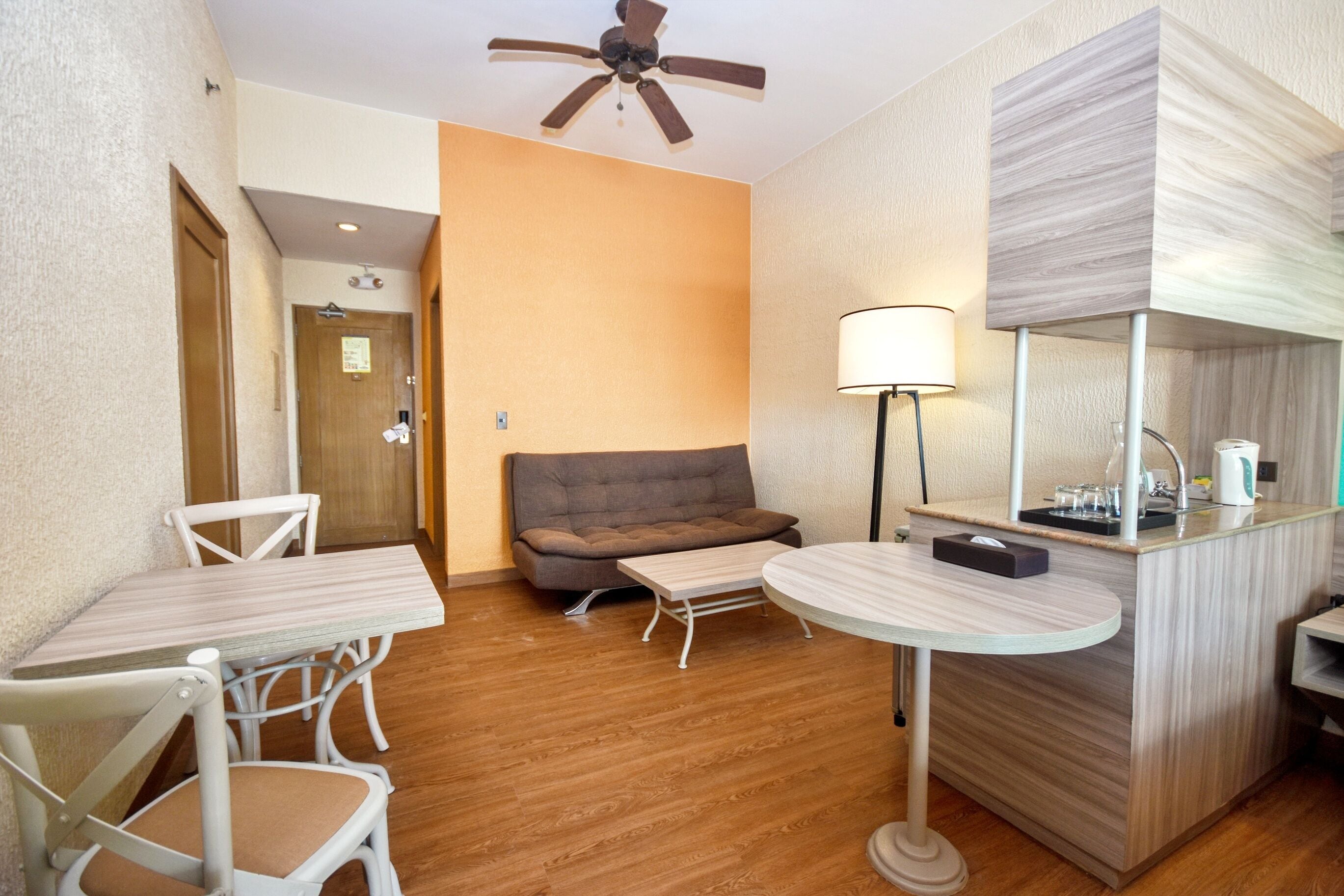 A hotel room with a wooden floor, a sofa, a small table, a round table, a mirror, and a ceiling fan.