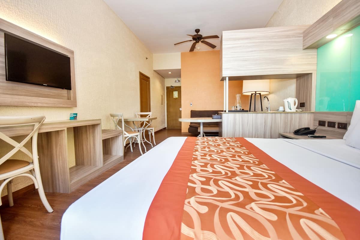 Habitación de hotel con cama, escritorio, silla, televisor, ventilador de techo y baño con espejo y lavamanos.