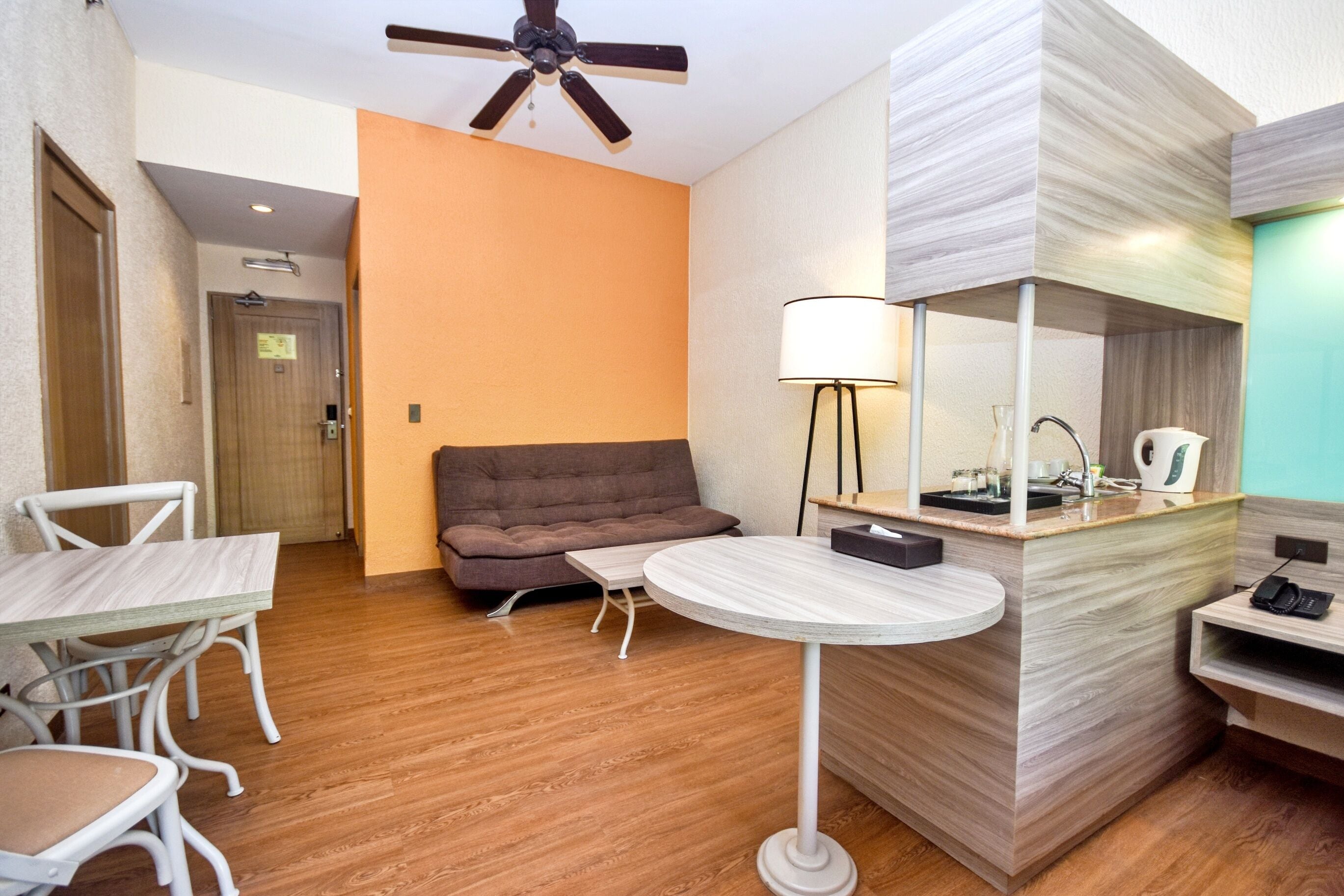 A modern hotel room with a wooden floor, a brown sofa, a round table, and a small desk with a lamp.