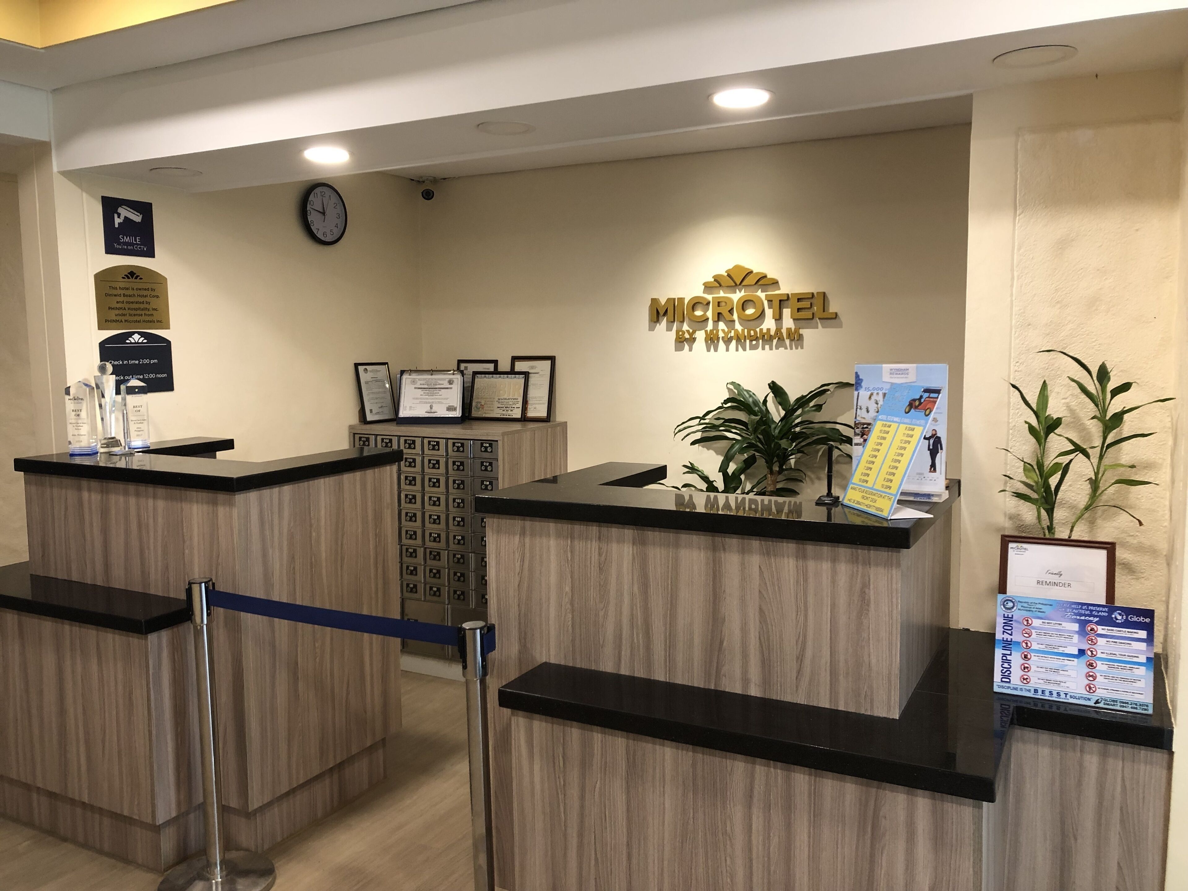 A hotel reception area with a reception desk, a clock, framed certificates, and a plant.