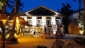 A two-story building with a balcony, surrounded by palm trees and outdoor seating.