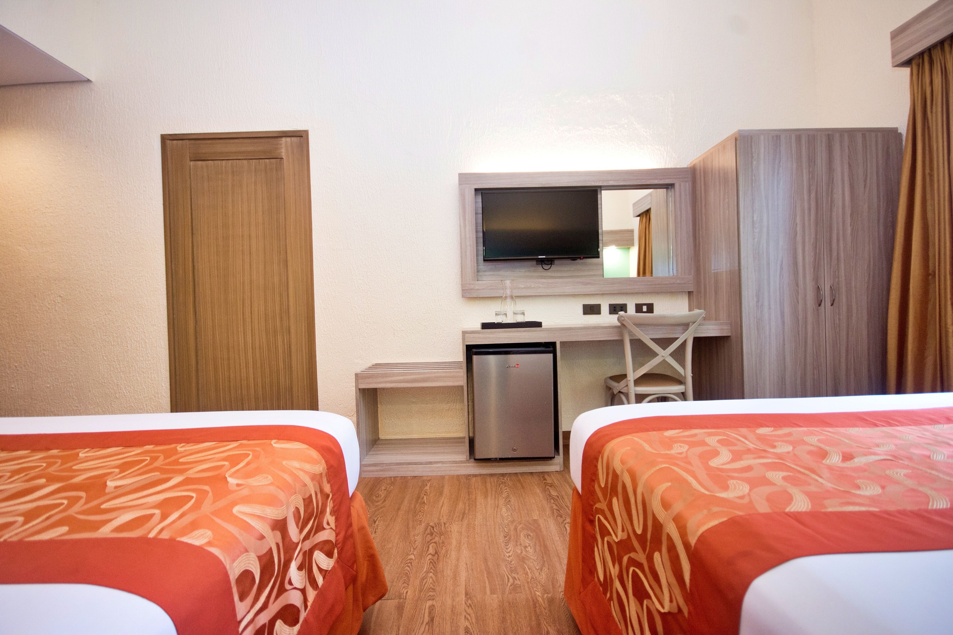 Une chambre d’hôtel avec deux lits, un bureau avec une télévision, un petit réfrigérateur et une armoire.