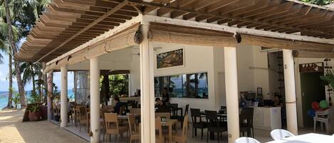 Un café al aire libre con mesas y sillas, una pérgola de madera y vistas al océano.