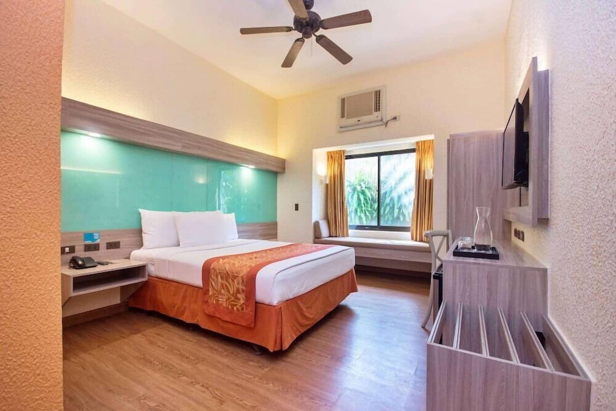 Una habitación de hotel con cama, mesita de noche, televisor y ventana con cortinas.