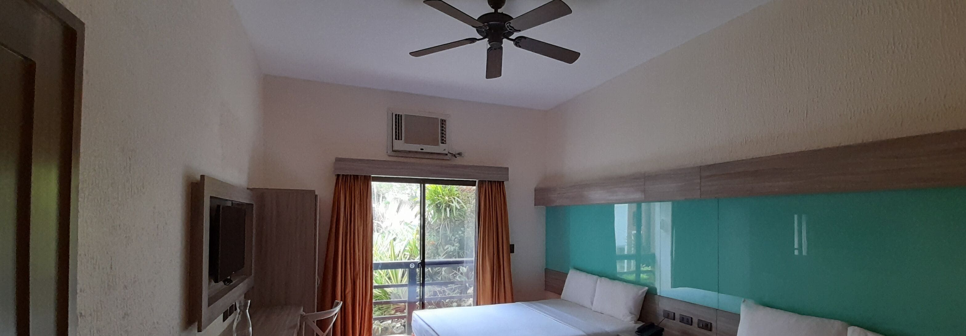 A hotel room with a bed, a ceiling fan, a TV, and a window with curtains.