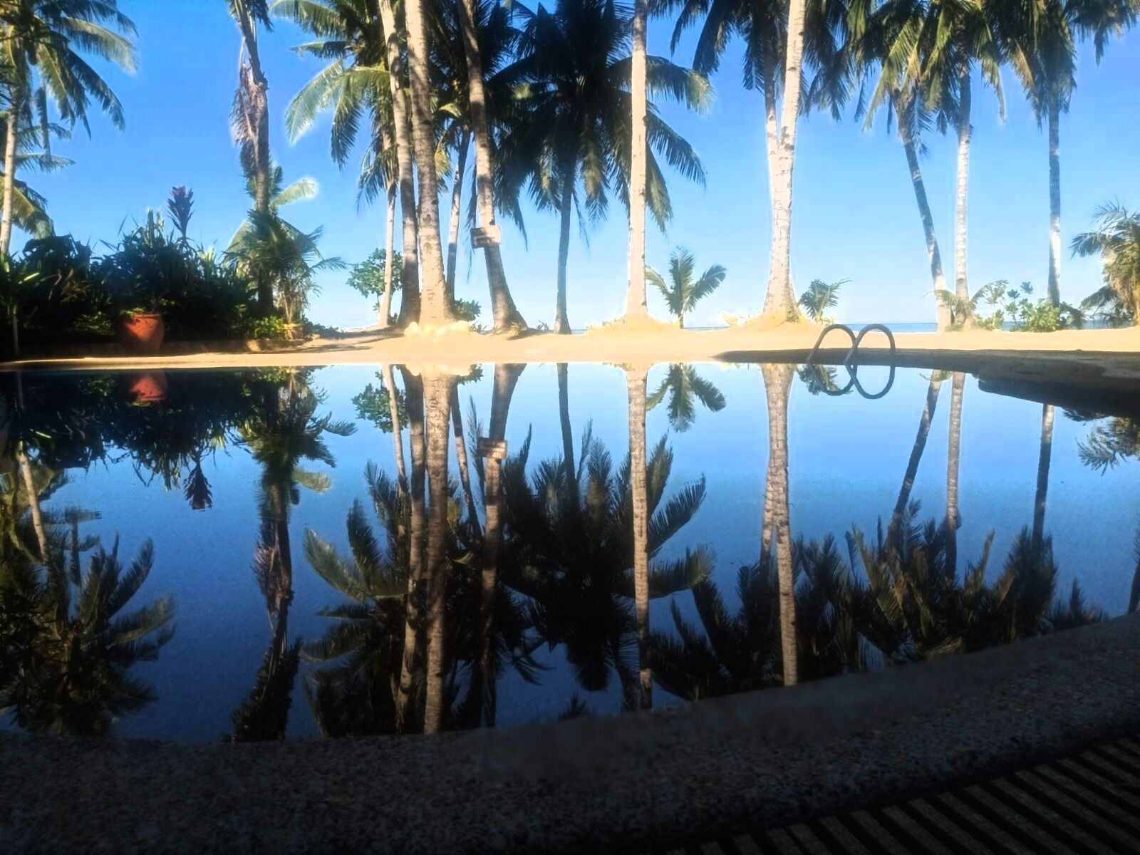 A tranquil pool area with palm trees reflected in the water, surrounded by a sandy beach and a clear blue sky.