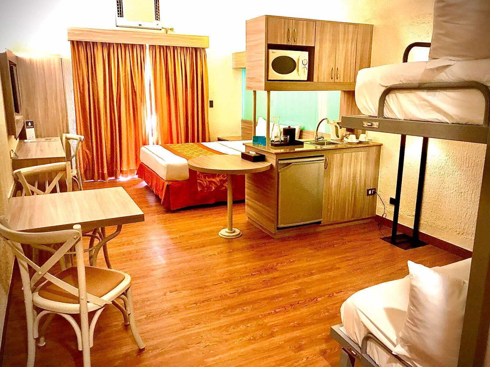 Habitació d'hotel senzilla amb un llit superposat, una cuineta petita, una zona de menjador amb taula i cadires, i una finestra gran amb cortines.
