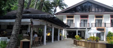 Un hotel con zona de estar al aire libre techada, palmeras y un edificio de dos pisos con balcones.