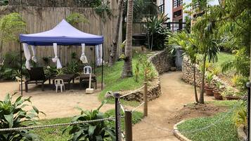 A covered outdoor seating area with chairs and a table, surrounded by greenery and a bamboo fence.