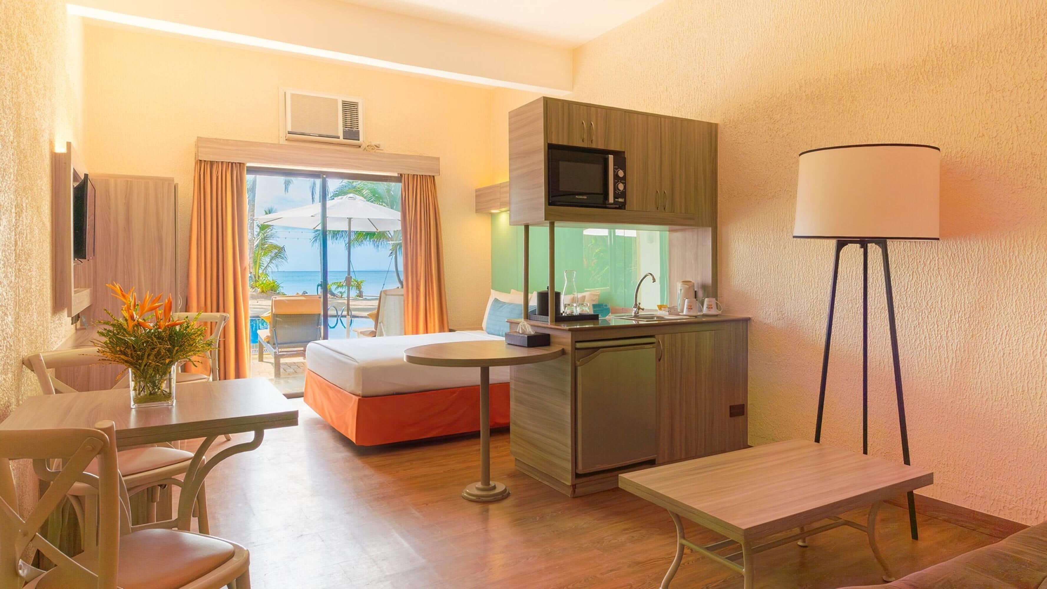 Habitación de hotel compacta con cama, pequeña kitchenette, mesa de comedor y vista al océano.
