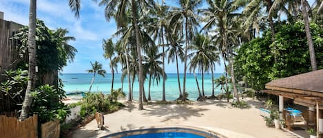 Zona de piscina con vista a la playa y al océano, rodeada de palmeras.