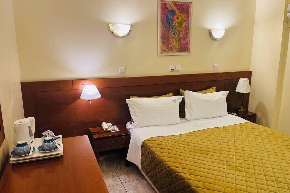 Una habitación de hotel con una cama, lámparas de noche, una mesita de noche, un teléfono, un cuadro en la pared y una mesa con una bandeja de tazas.