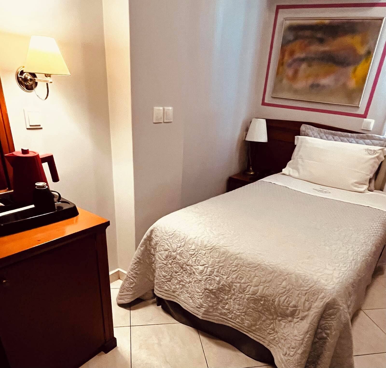 Una habitación de hotel con una cama, una mesita de noche, una lámpara, una cafetera y un cuadro en la pared.