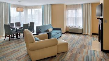A hotel room with a sofa, a dining table with chairs, a TV, and large windows with curtains.