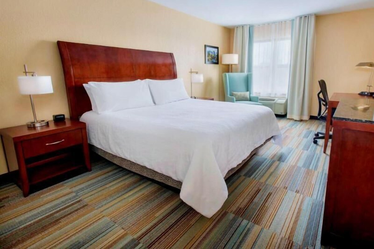 Quarto de hotel com uma cama grande, escrivaninha, cadeira e janela com cortinas.