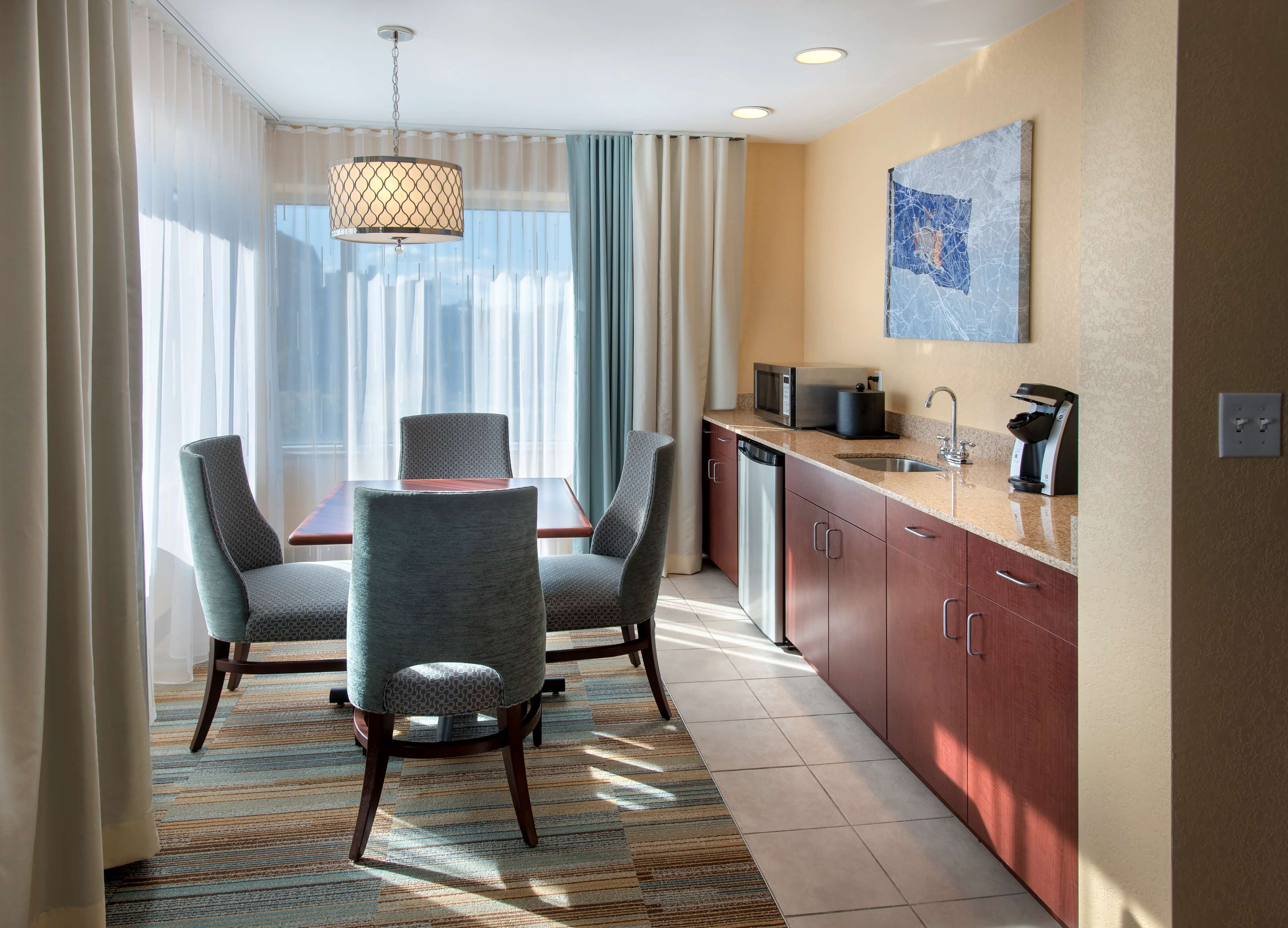 A hotel room with a dining table, four chairs, a kitchen area with a microwave, coffee maker, and a sink.