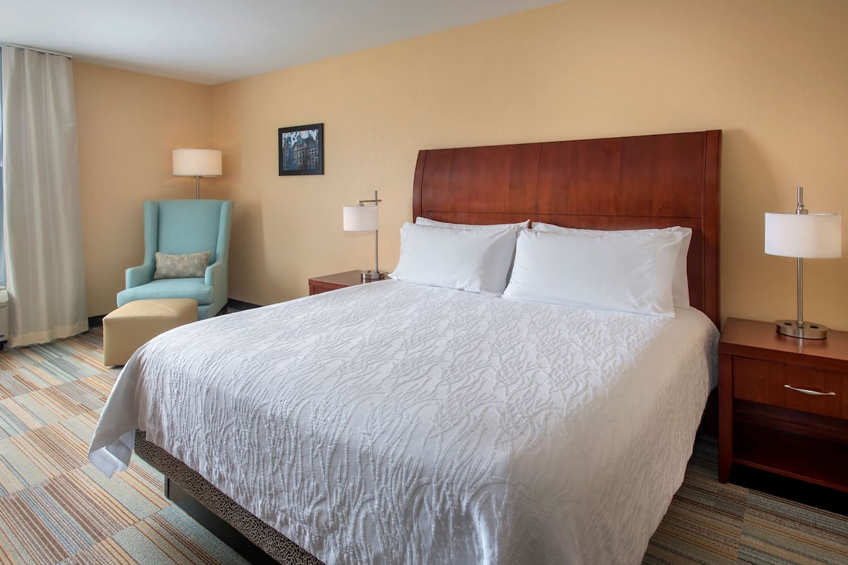 A hotel room with a large bed, a chair, two bedside tables with lamps, and a window with curtains.