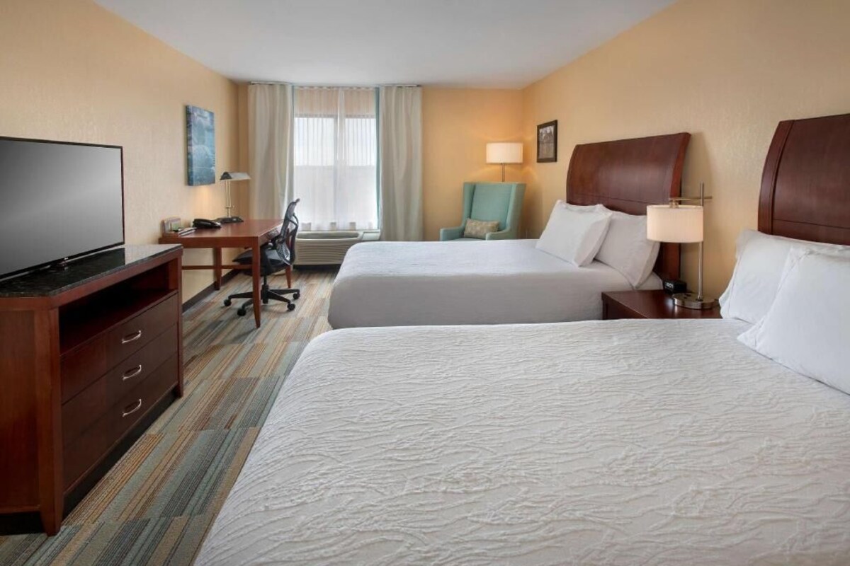 Quarto de hotel com duas camas, uma mesa com televisão, uma cadeira, abajur e janela com cortinas.