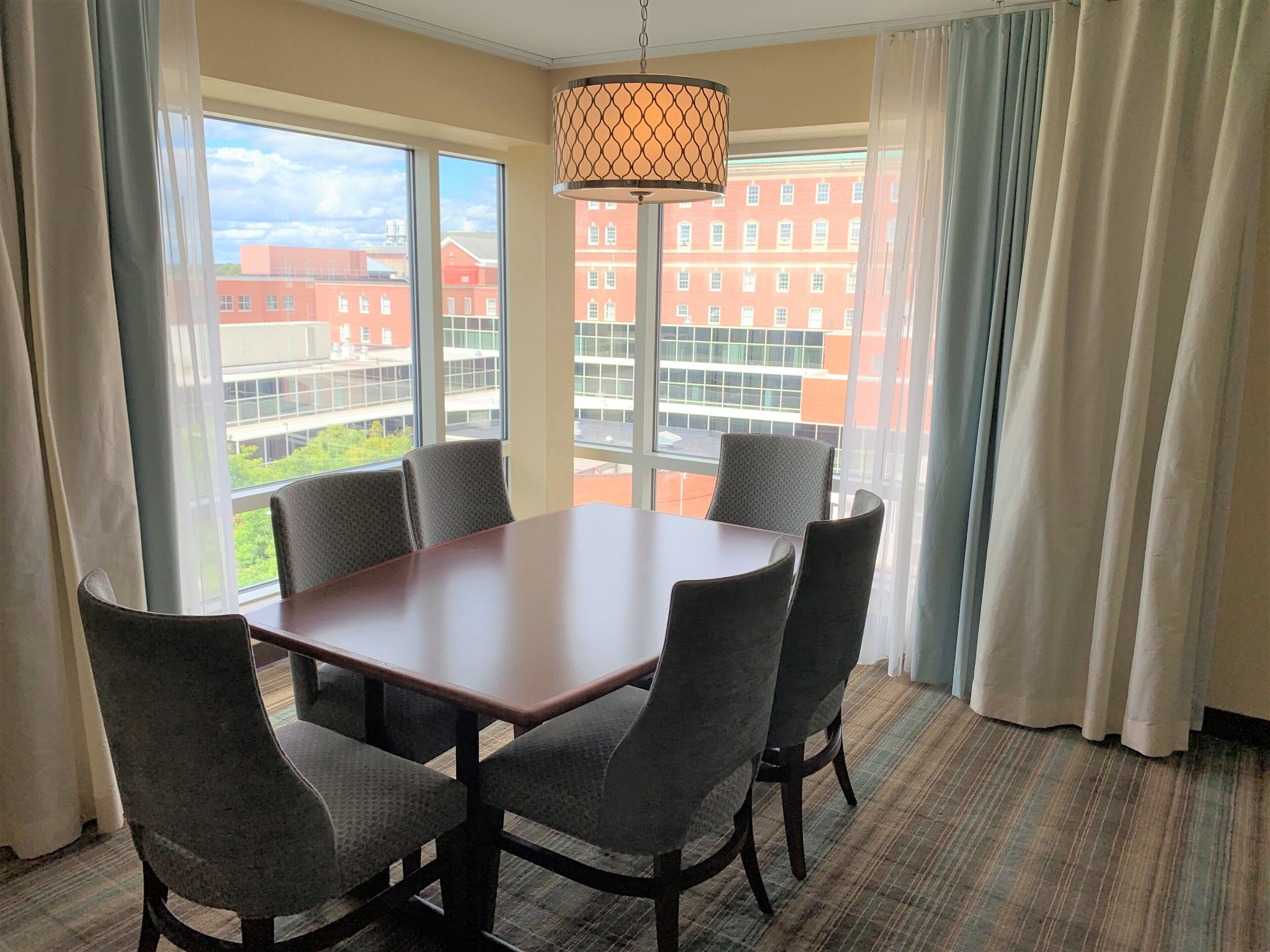 A conference room with a large table and six chairs, large windows with curtains, and a hanging lamp.