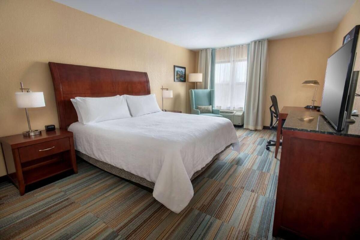 A hotel room with a large bed, a desk, a chair, a TV, and a lamp.